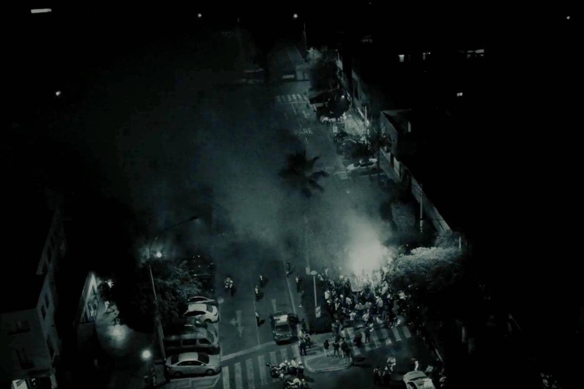 Pemandangan malam dari atas memperlihatkan kerumunan orang di jalan dengan asap tebal dan cahaya terang di tengah suasana gelap.