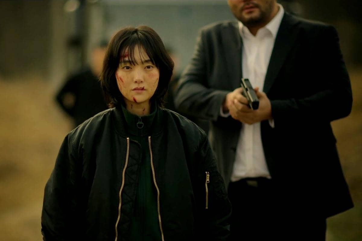 Seorang wanita dengan wajah berdarah berdiri di depan pria bersetelan jas yang memegang pistol dalam adegan drama A Shop for Killers 2.