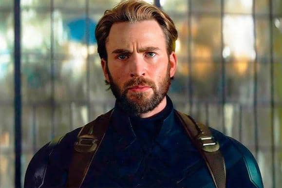 Seorang pria berjanggut mengenakan kostum superhero berwarna gelap dengan latar belakang kaca buram dalam adegan film aksi.