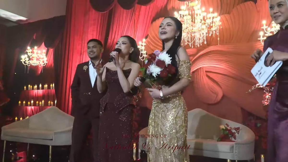 Suasana resepsi pernikahan dengan dekorasi merah dan emas, pasangan pengantin berdiri di panggung bersama tamu yang memegang mikrofon dan bunga.