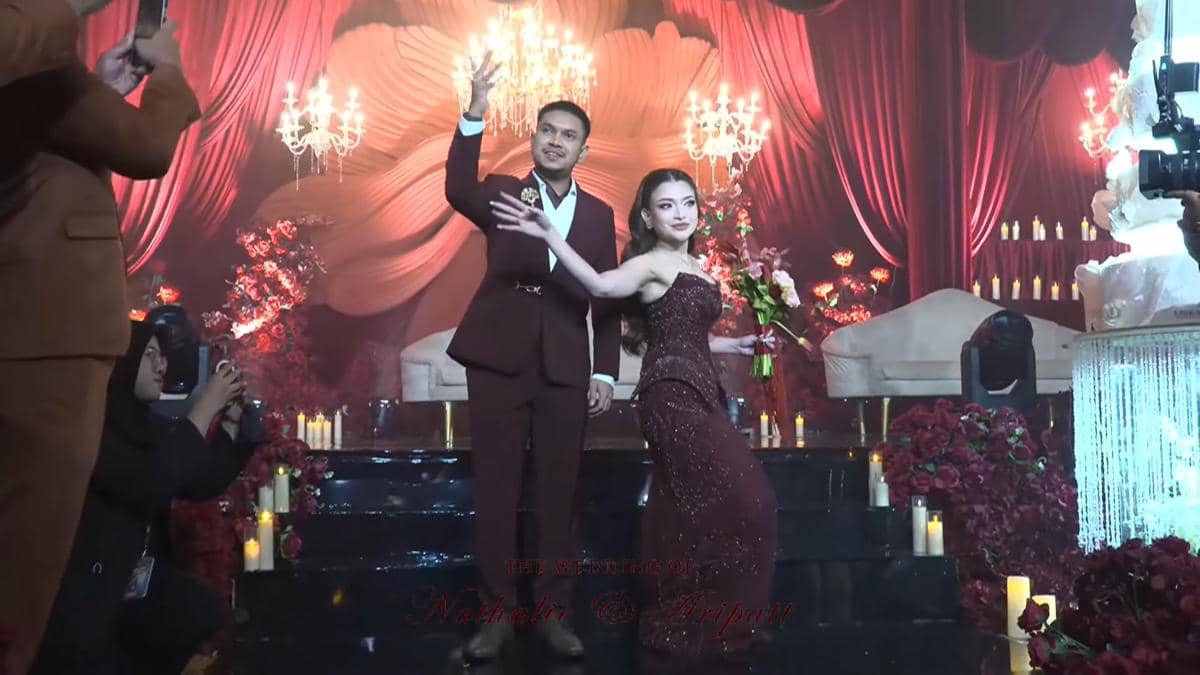 Pasangan pengantin mengenakan busana serasi warna marun berpose di panggung resepsi dengan dekorasi bunga merah dan lampu gantung mewah.
