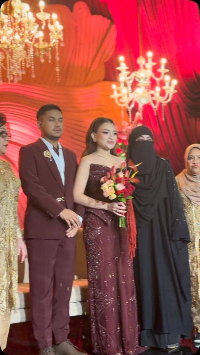 Pasangan pengantin berbusana marun berpose bersama keluarga di panggung resepsi dengan dekorasi merah dan lampu gantung mewah.