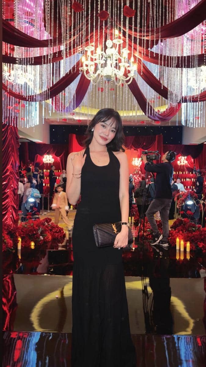 Seorang wanita bergaun hitam elegan berpose di area resepsi pernikahan dengan dekorasi merah dan lampu gantung mewah.