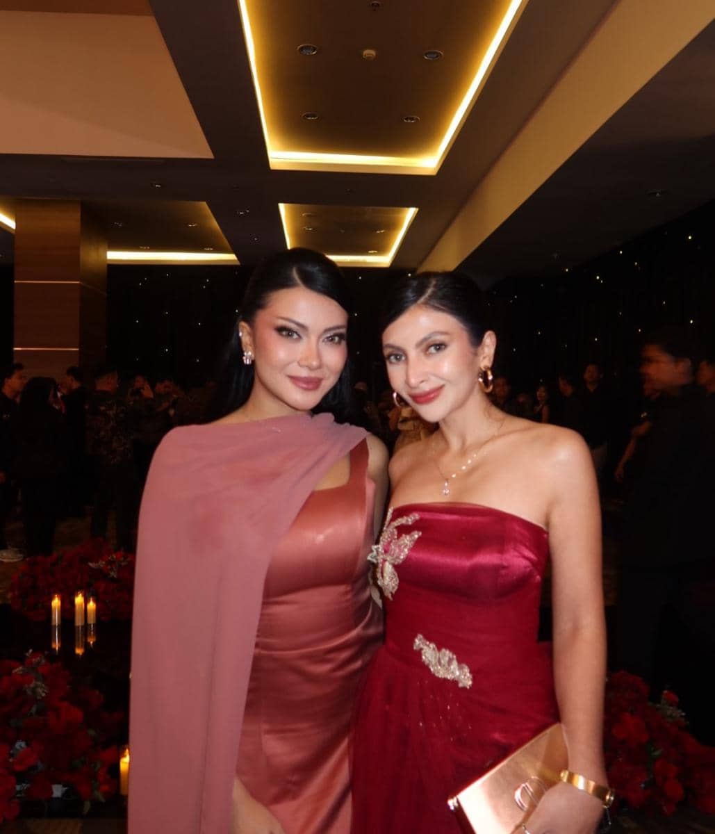 Dua tamu wanita berpose mengenakan gaun elegan berwarna merah dan merah muda di resepsi pernikahan dengan latar dekorasi mewah.
