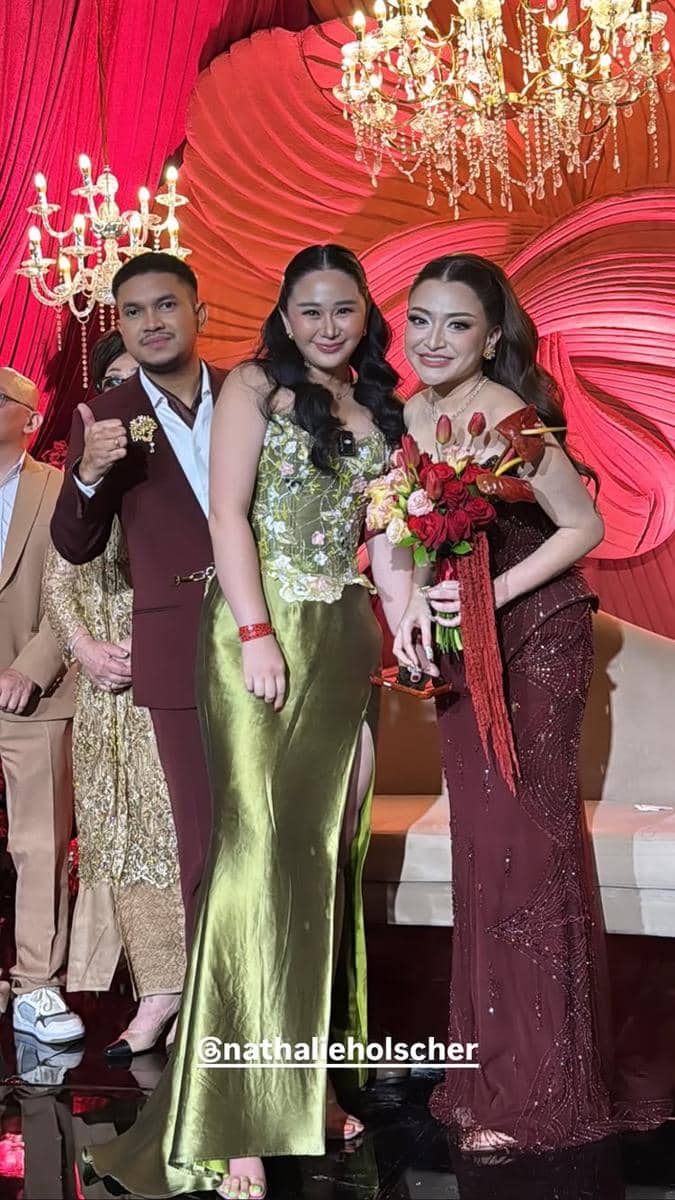 Beberapa tamu berpose bersama pasangan pengantin di resepsi pernikahan dengan dekorasi merah elegan dan lampu gantung mewah.