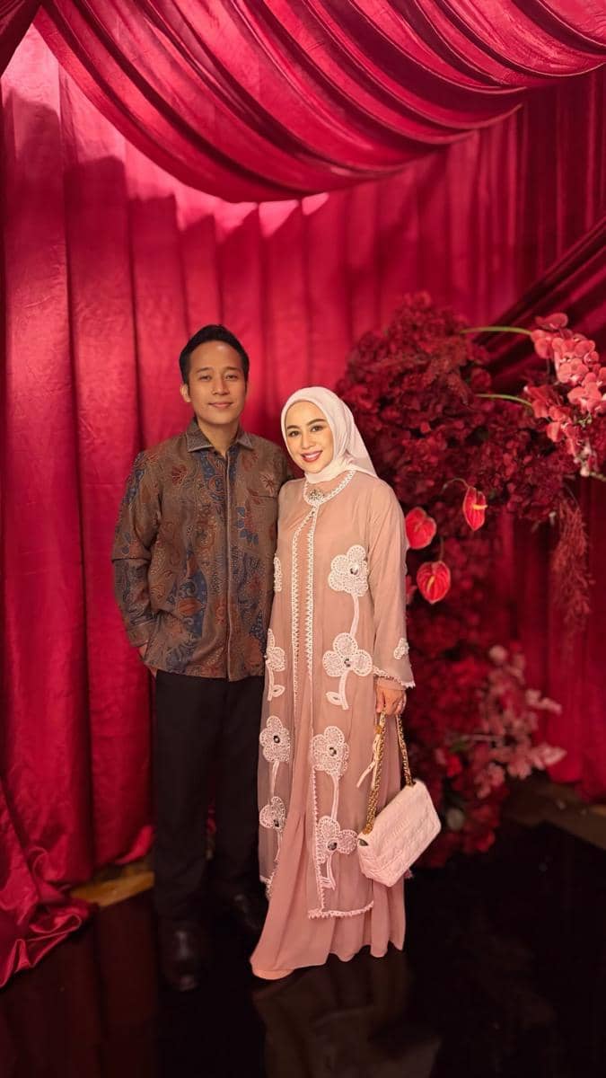 Sepasang tamu berpose di resepsi pernikahan dengan latar dekorasi merah marun dan bunga hias berwarna senada.