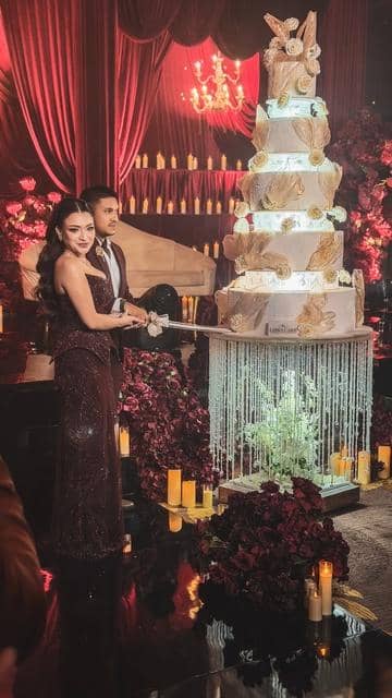 Pasangan pengantin mengenakan busana formal elegan sedang memotong kue pernikahan bertingkat tinggi di ruang resepsi mewah dengan dekorasi bunga dan lilin.