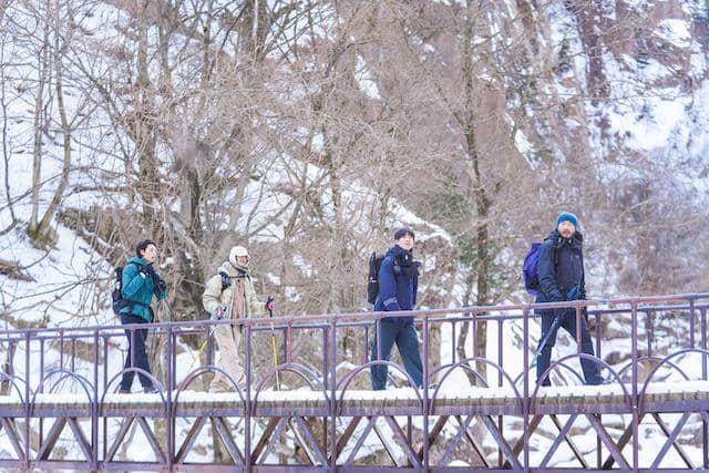 Empat orang pendaki berjalan di atas jembatan kayu saat musim dingin dengan latar pepohonan bersalju di belakang mereka.