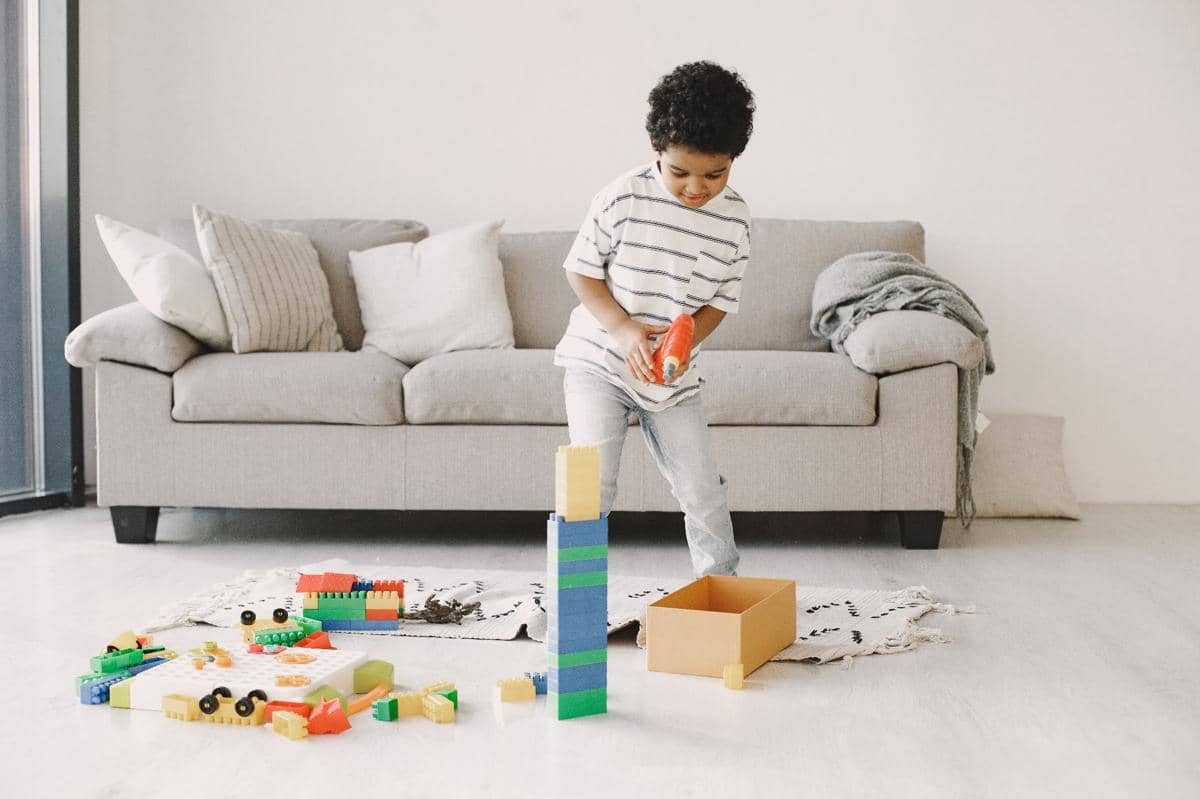 Seorang anak kecil bermain building blocks berwarna-warni di ruang tamu dengan sofa abu-abu di latar belakang.