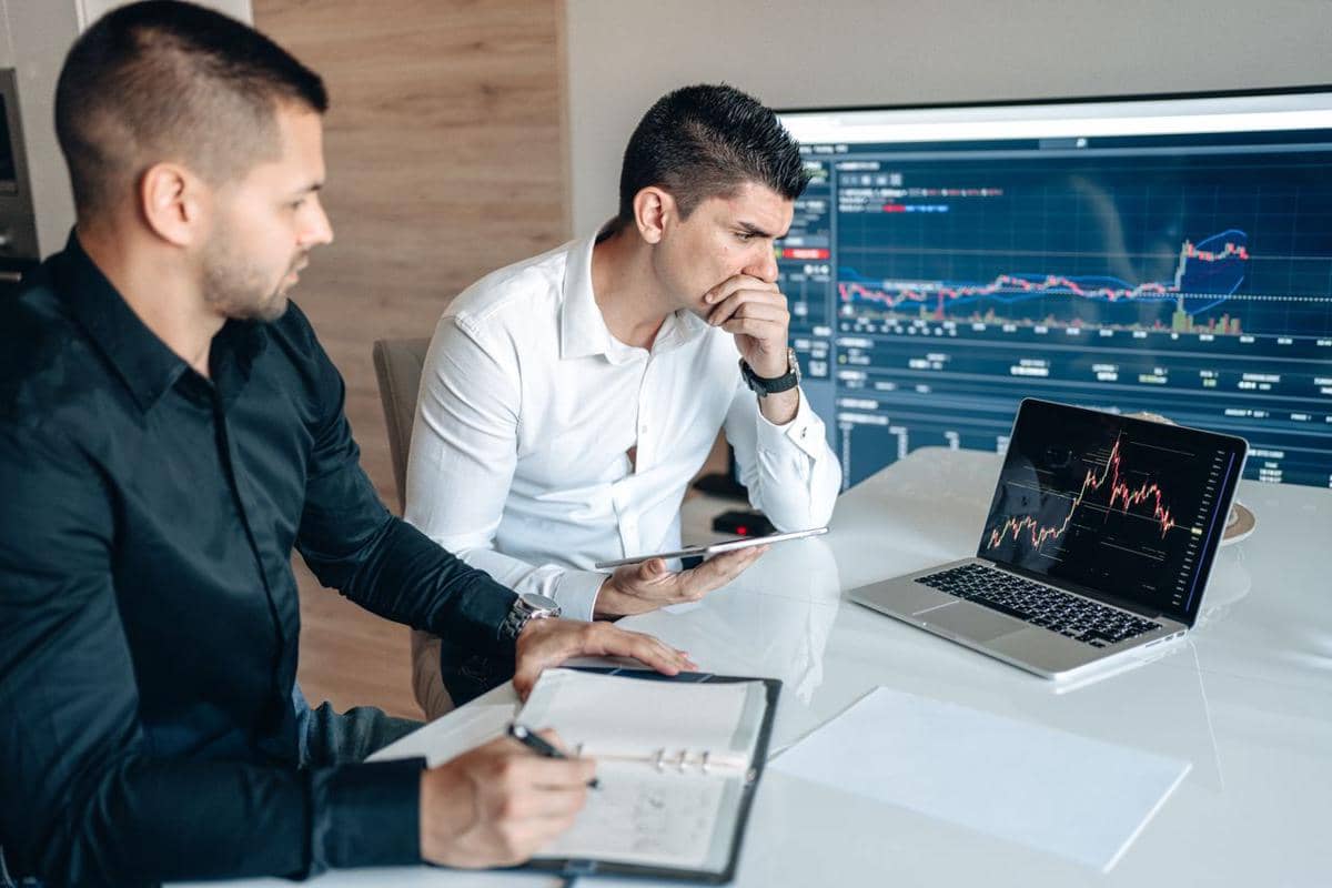 ilustrasi dua pengusaha menganalisis data pasar saham di laptop dan tablet