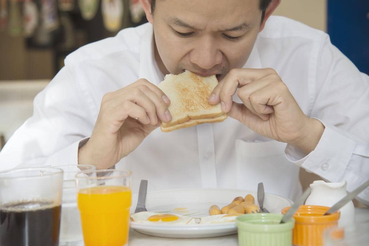 Ilustrasi seseorang mengenakan pakaian santai sedang menikmati sepotong roti sambil duduk di dalam ruangan.