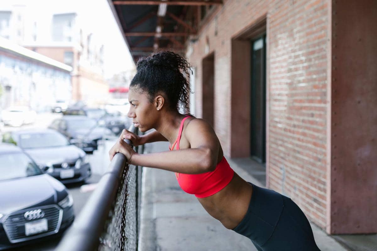 Seorang perempuan mengenakan pakaian olahraga melakukan wall push-up dengan kedua tangan bertumpu pada dinding putih.