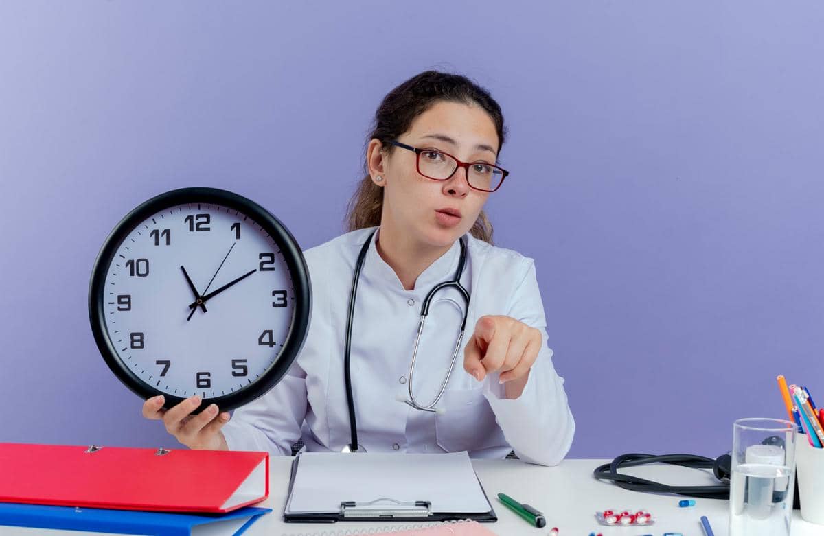 Seorang dokter memegang jam dinding sambil menunjuk ke arah kamera di ruang kerja