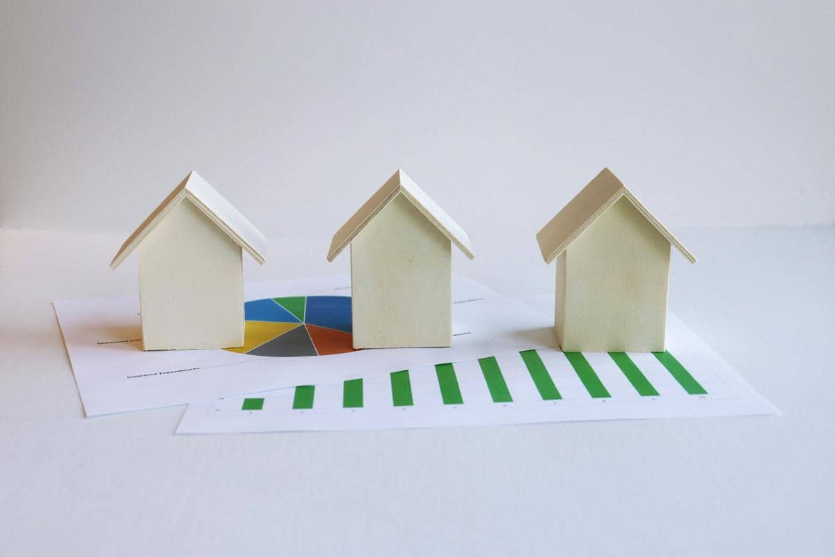 Tiga miniatur rumah berdiri di atas dokumen berisi grafik keuangan