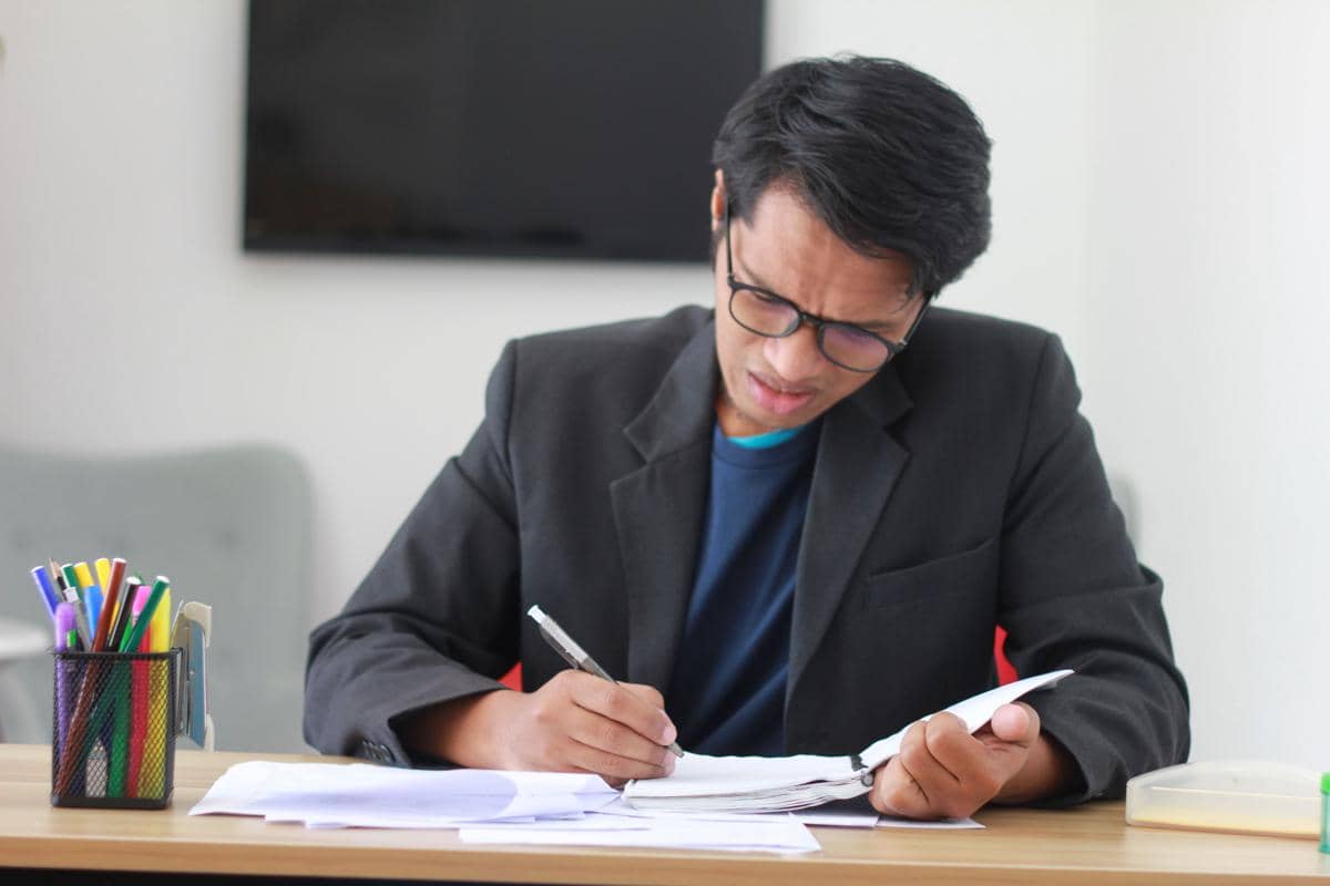Seorang pria berkacamata menulis dan memeriksa sejumlah dokumen di meja kerja