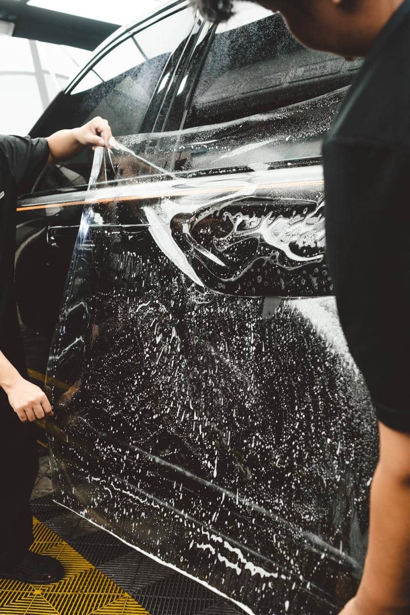 Seorang teknisi memasang paint protection film (PPF) pada permukaan kendaraan dengan teliti di area kerja.