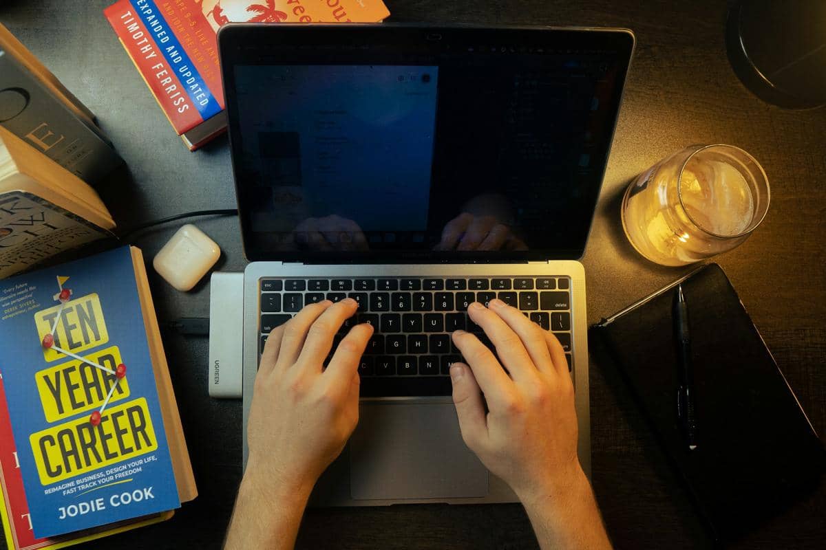 Seseorang mengetik di laptop di antara buku dan catatan kerja.