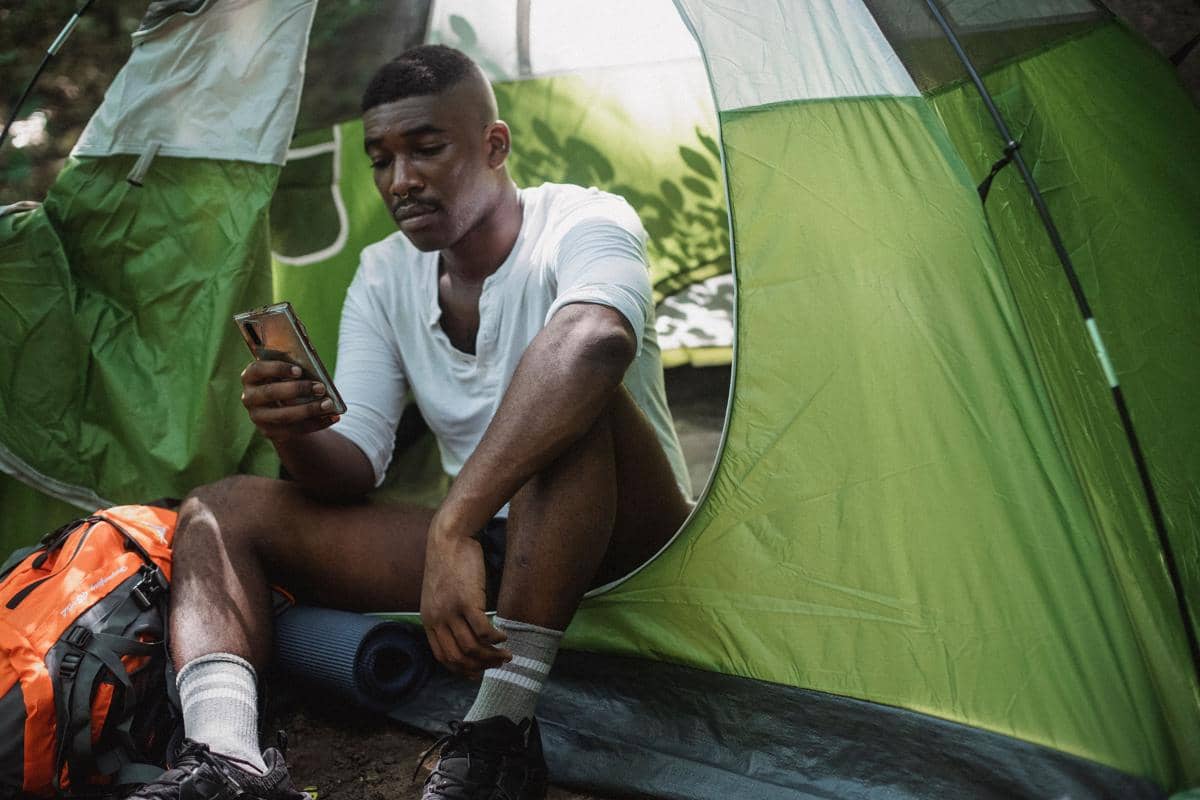 Seorang pria memeriksa ponsel sambil duduk di depan tenda camping