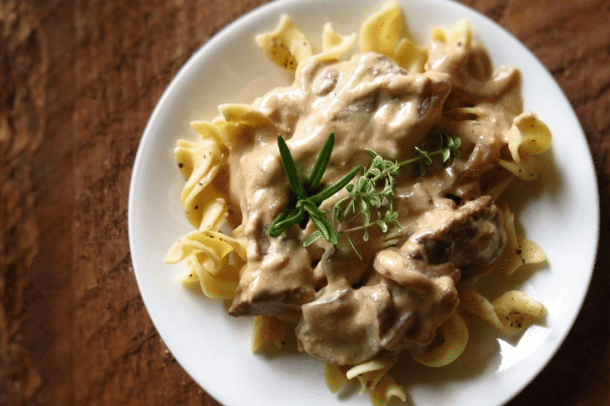 Sajian beef stroganoff di atas piring dengan saus krim berwarna cokelat muda dan potongan daging sapi.