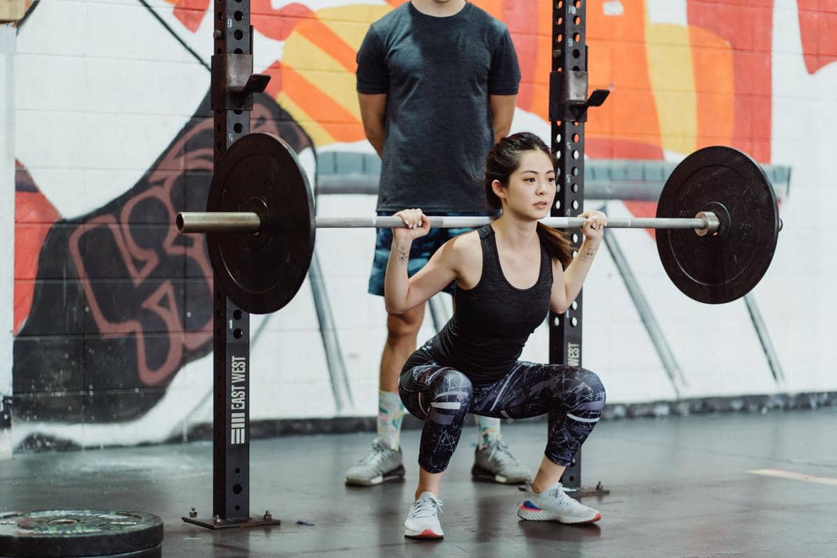 Seorang wanita melakukan gerakan barbell squat dengan barbel di pundak sebagai latihan compound di area gym.