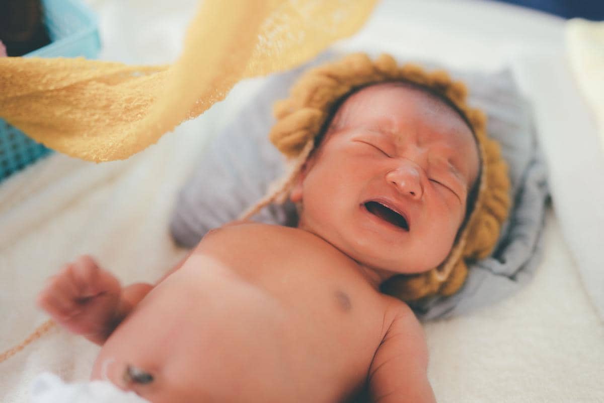 Seorang bayi menangis sambil berbaring di atas tempat tidur, dengan tangan terangkat dan wajah tampak sedih.