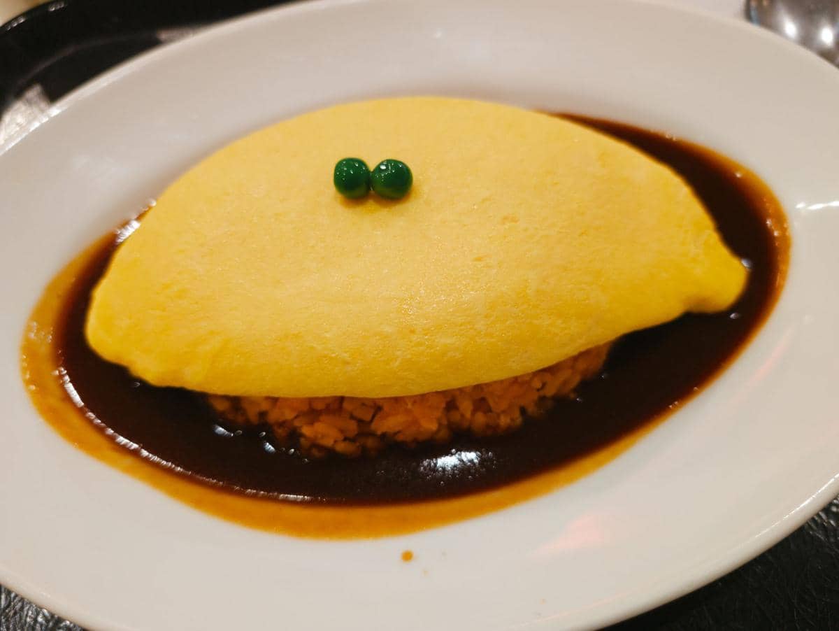 Sepiring omurice berupa nasi goreng yang dibungkus telur dadar, disajikan dengan saus di atas piring putih.