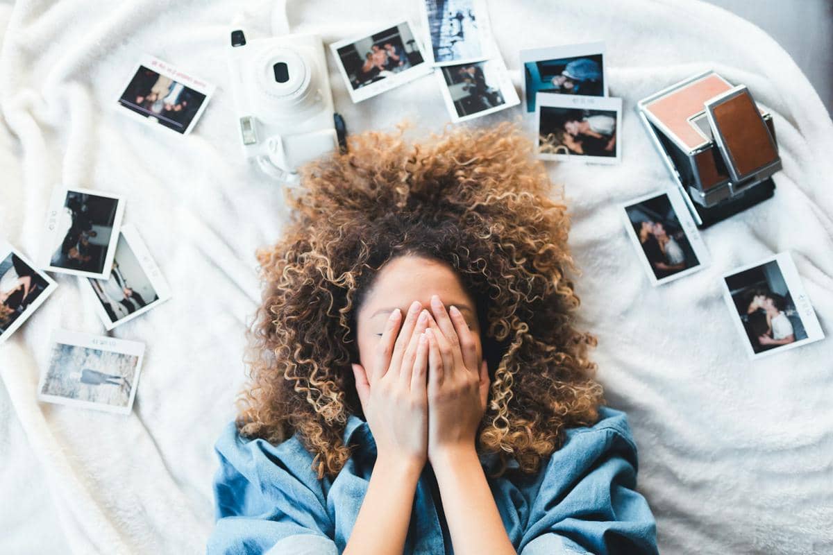Seseorang menutupi wajahnya sambil berbaring di antara foto-foto cetak dan kamera, menggambarkan perasaan cemas dan refleksi diri.