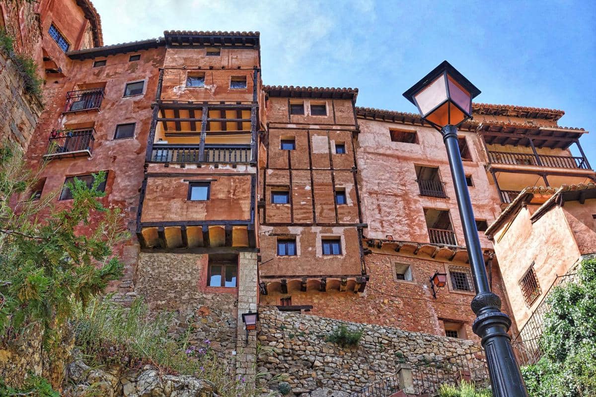 Deretan rumah berwarna terakota di Kota AlbarracÃn, Spanyol, berdiri di lereng berbatu dengan perbukitan hijau di kejauhan.