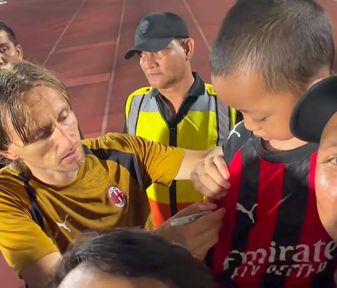 Oki Rengga dan Arthur berfoto bersama pemain AC Milan di lapangan latihan dengan suasana tim di belakang mereka.