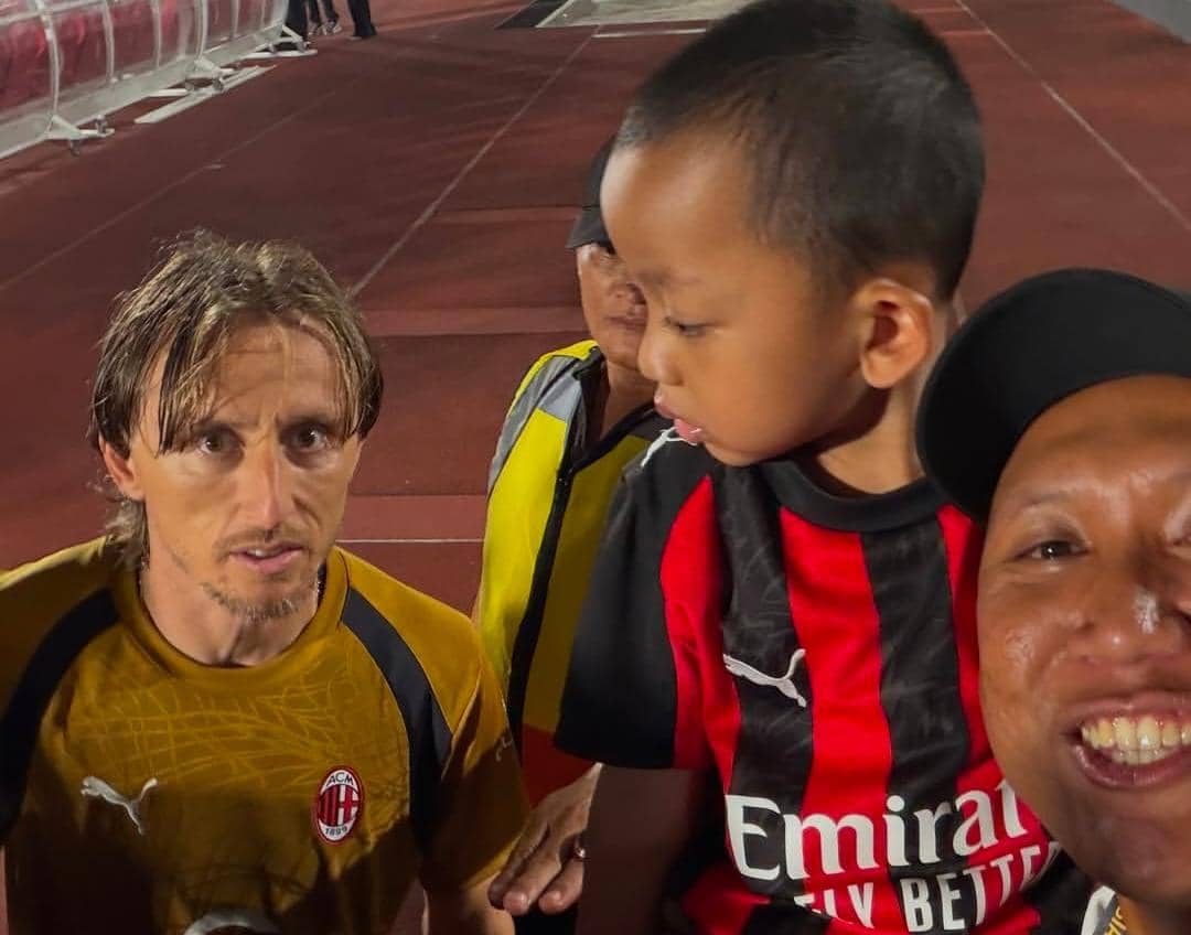 Oki Rengga dan Arthur berpose bersama pemain AC Milan di lapangan latihan klub Italia dengan latar rumput hijau.