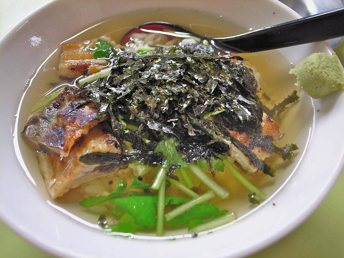 Ilustrasi semangkuk ochazuke, hidangan nasi Jepang dengan kuah teh dan aneka pelengkap di atasnya.