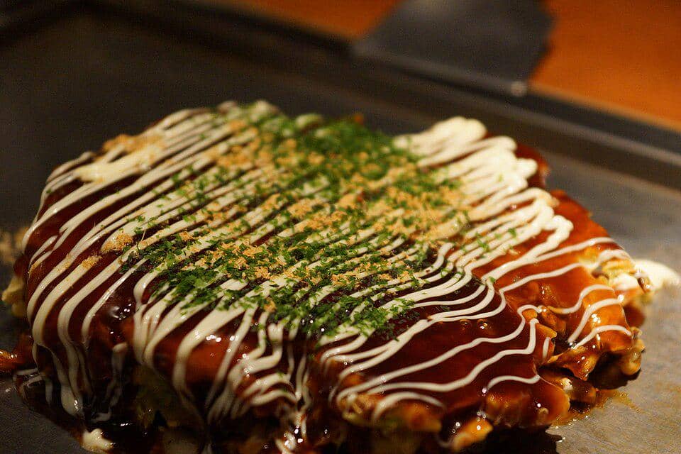 Ilustrasi hidangan okonomiyaki khas Jepang tersaji di atas piring, dengan beragam topping di permukaannya.