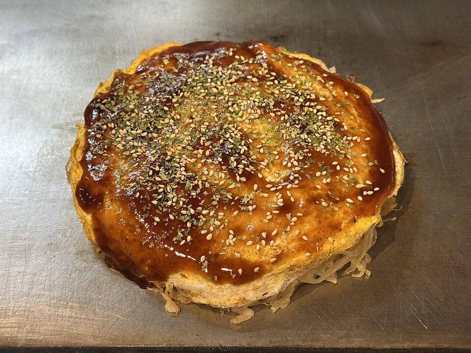 Sepiring okonomiyaki gaya Hiroshima dengan lapisan mi, telur, kol, dan saus cokelat, disajikan di atas piring putih.