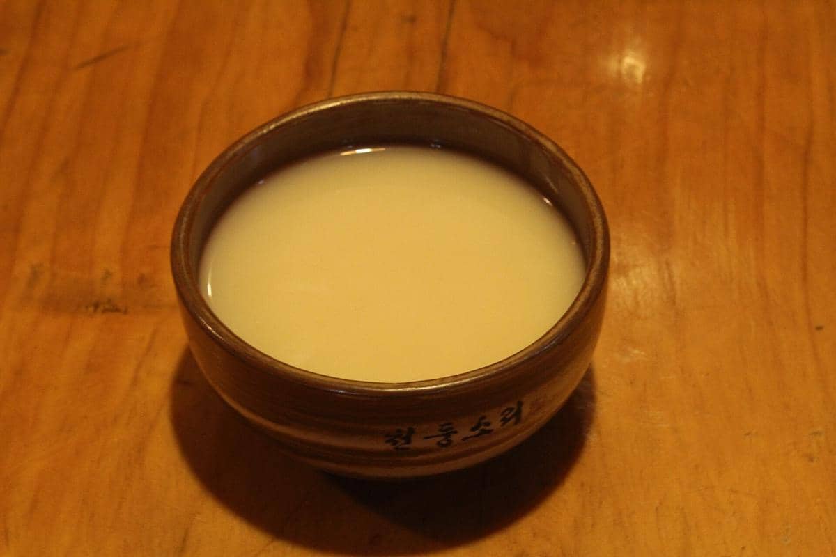 Ilustrasi makgeolli, minuman beras fermentasi khas Korea berwarna putih susu yang disajikan dalam mangkuk dan botol.