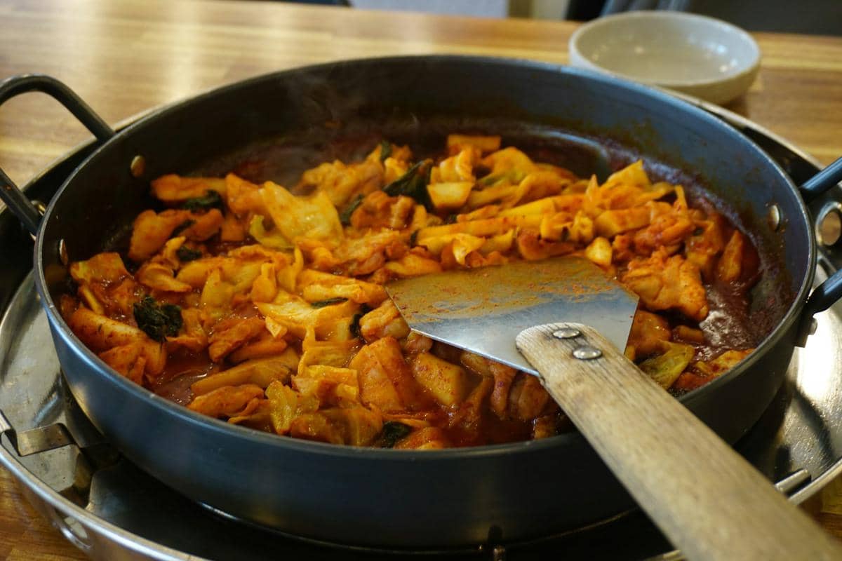 Ilustrasi proses membuat Korean zicoba chicken, hidangan ayam Korea berbumbu pedas yang disajikan di atas meja.