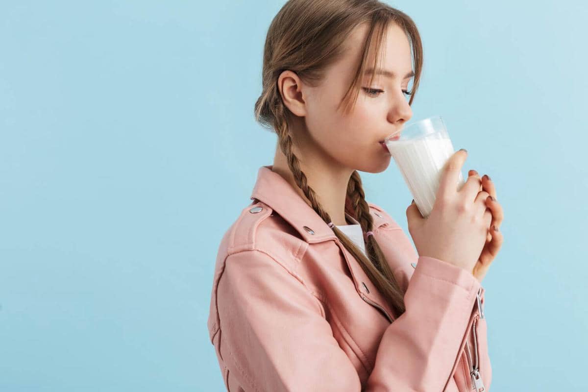 Ilustrasi seorang perempuan minum segelas susu putih sambil duduk di meja, dengan latar dapur bernuansa biru.