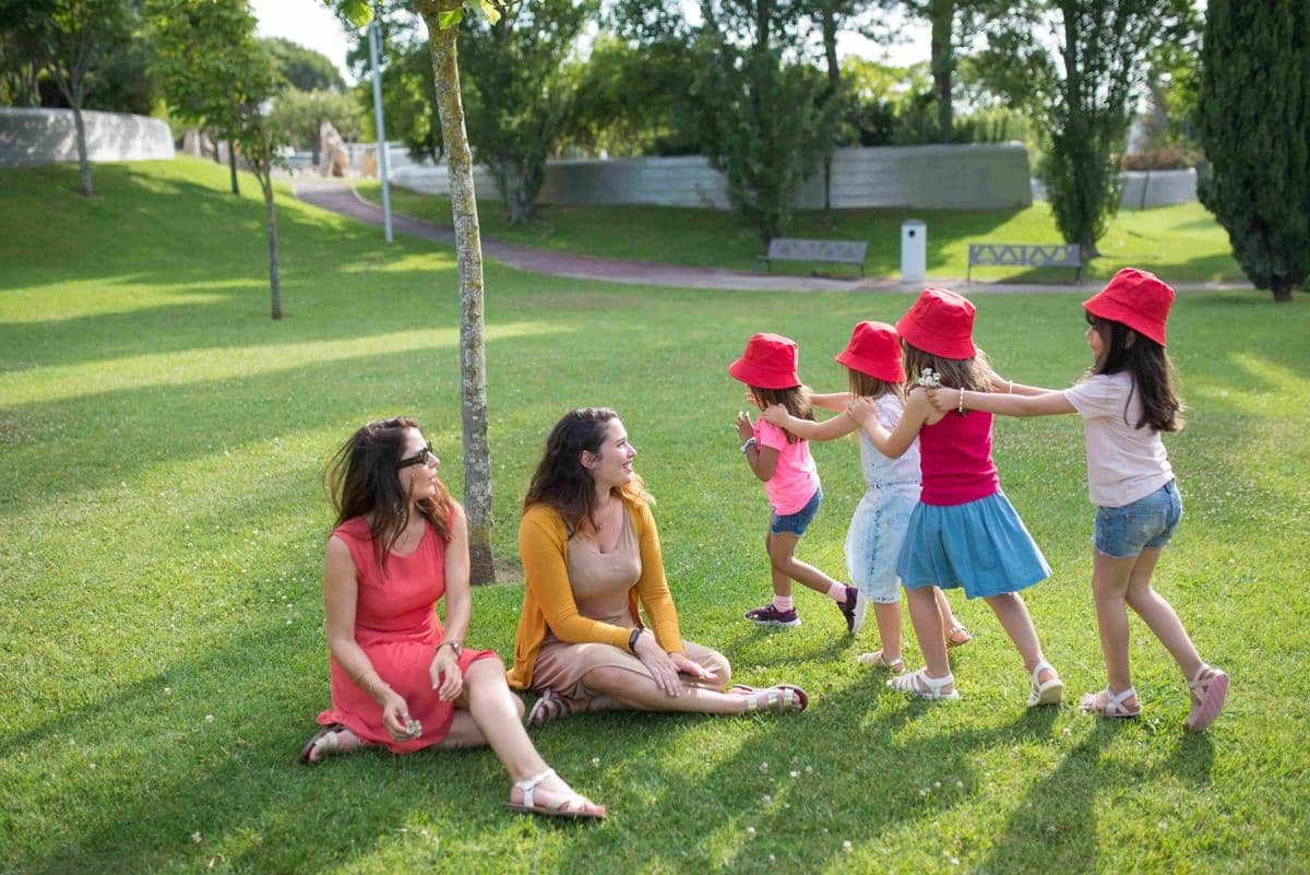 Seorang dewasa mengamati anak-anak yang sedang bermain bersama di taman dari kejauhan dengan penuh perhatian.