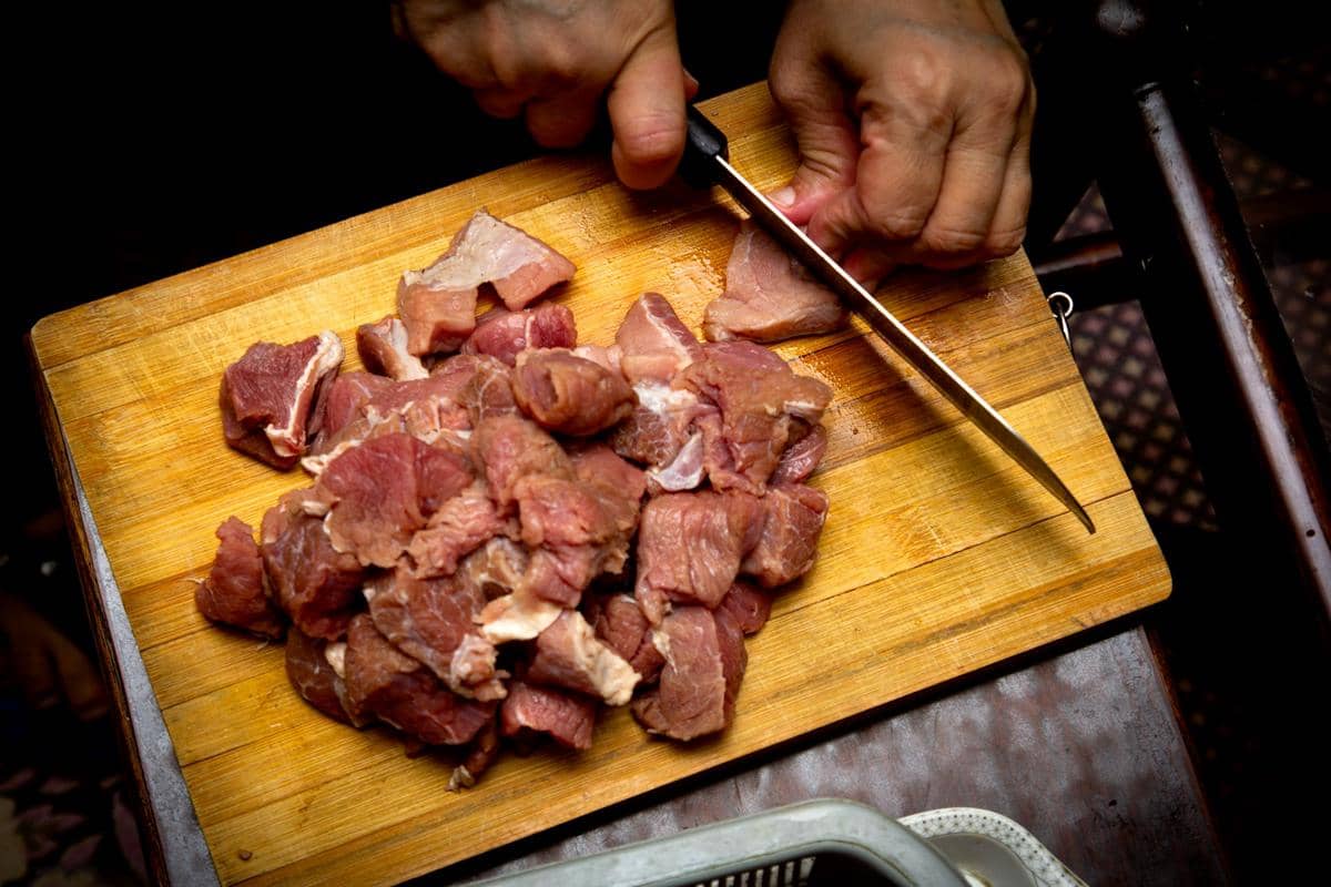 Tangan memotong daging mentah di atas talenan kayu menggunakan pisau dapur berbilah lebar di meja.