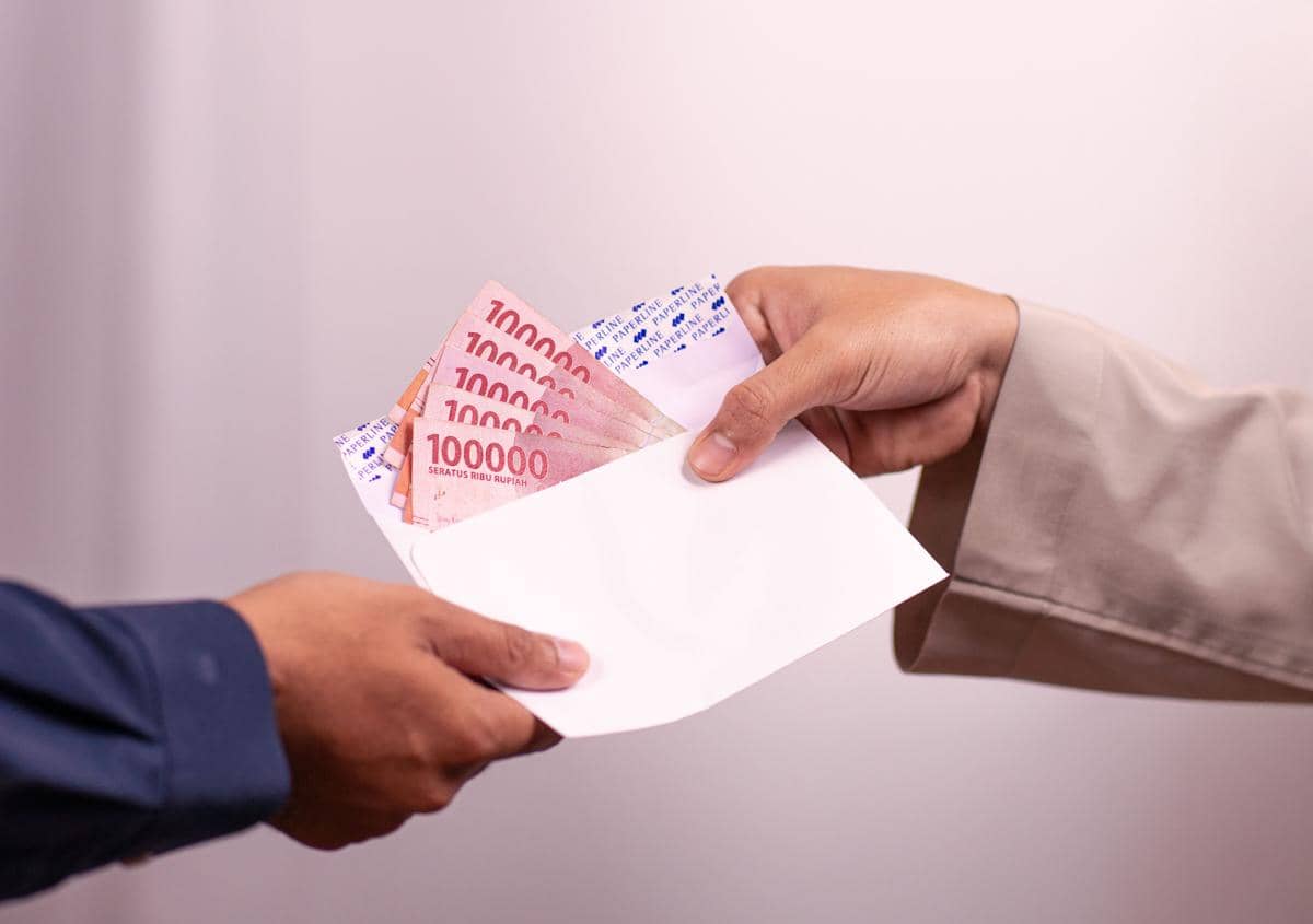 Dua orang saling menyerahkan amplop putih berisi uang lembaran Rupiah merah 100 ribu