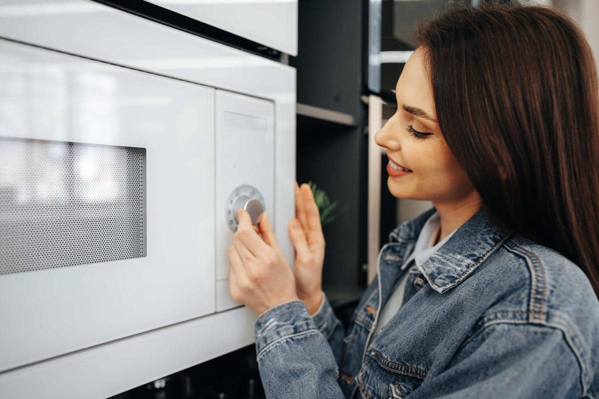 Seorang perempuan memeriksa microwave oven berwarna hitam di rak toko elektronik sebagai ilustrasi mematikan oven.