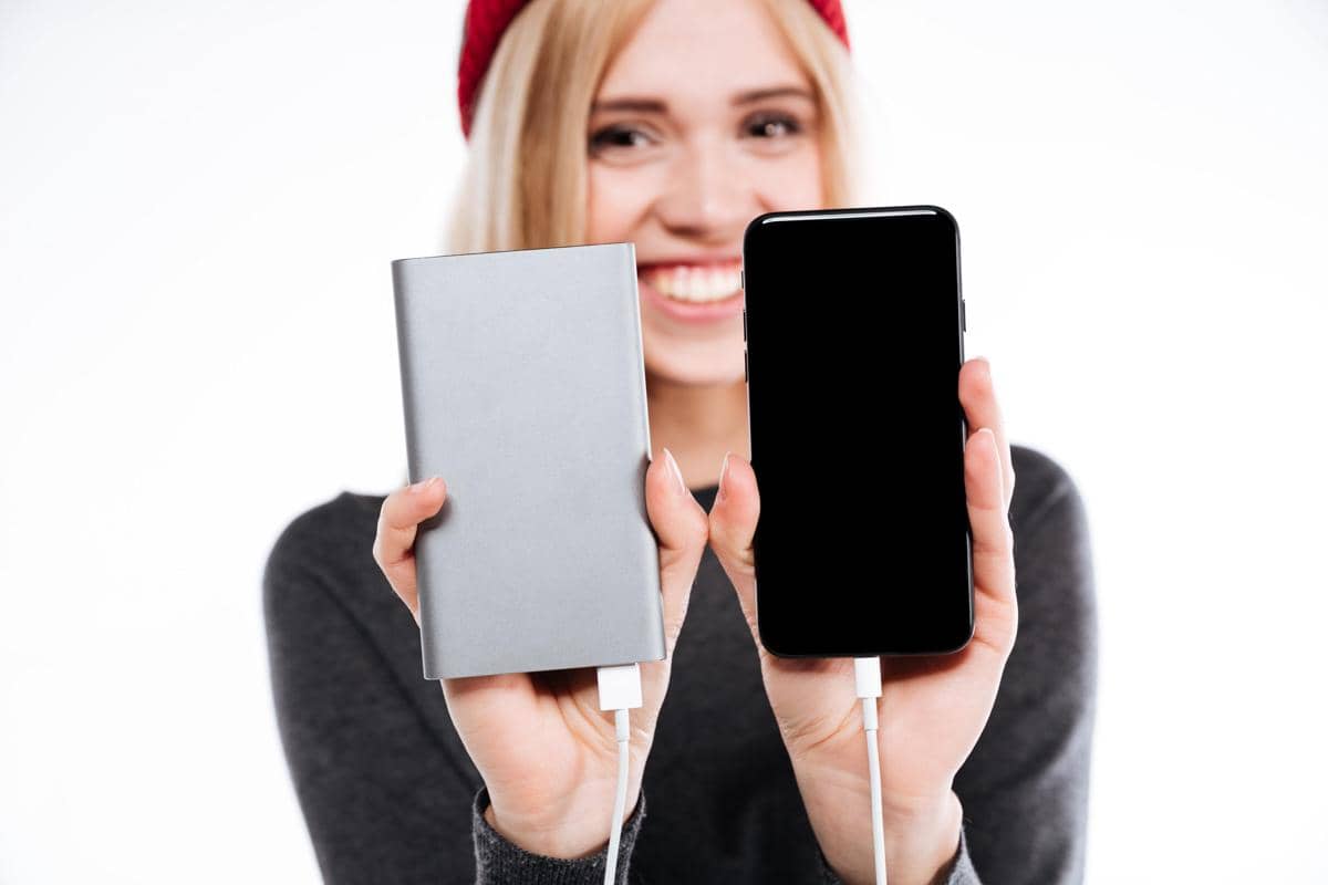 Seorang perempuan memegang ponsel dan powerbank putih, menggambarkan penggunaan pengisi daya portabel saat bepergian.