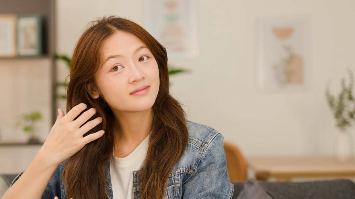 Perempuan muda Asia mengenakan jaket denim merapikan rambut panjangnya dengan tangan di dalam rumah yang terang.