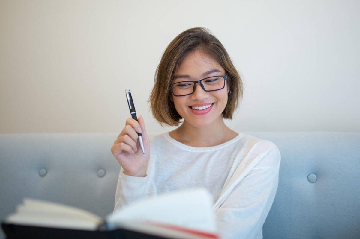 Seorang perempuan tersenyum duduk di sofa sambil membaca buku dan mengangkat pulpen di ruang tamu rumah.