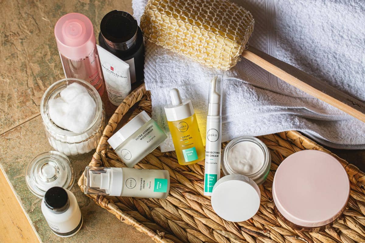 Sebuah keranjang anyaman cokelat berisi berbagai macam botol serum, toner, wadah krim wajah, kapas pembersih, dan sikat badan kayu di atas kain putih.
