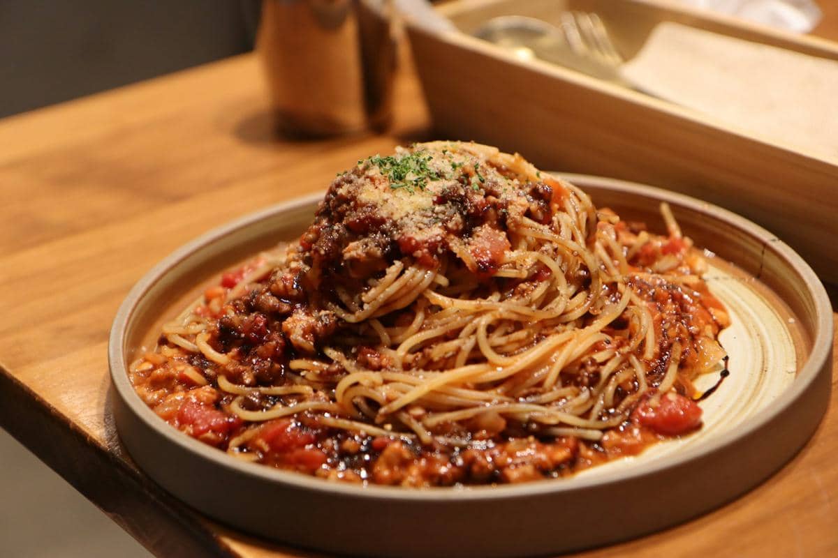Pasta alla trapanese tersaji dalam mangkuk putih dengan saus merah, tomat ceri, dan daun basil di atas meja.