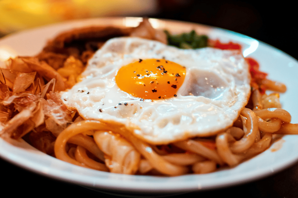 Semangkuk udon butter masala gochujang dengan mi tebal berbalut saus merah krem dan taburan daun bawang.