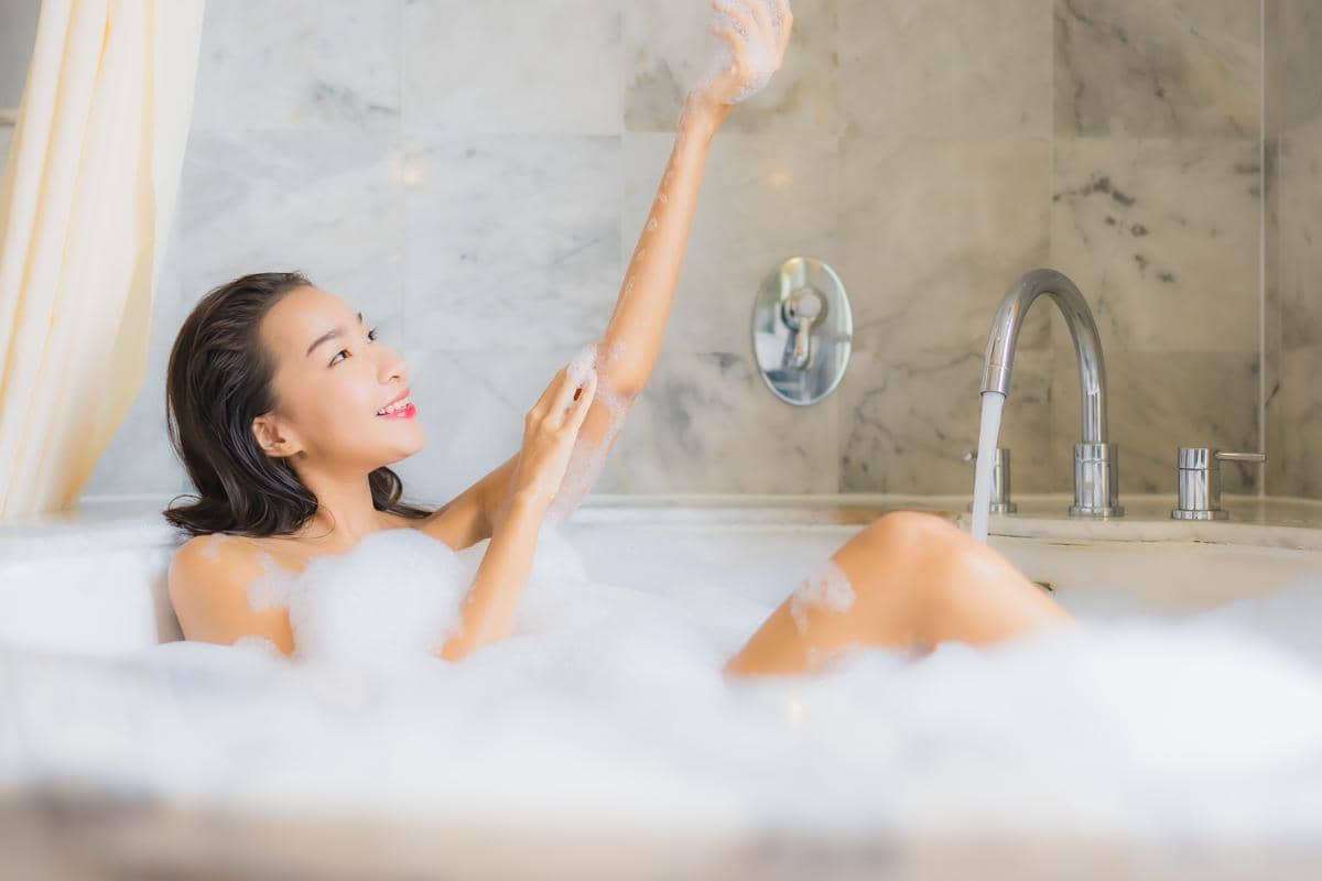 Seorang perempuan muda Asia berendam santai di bak mandi dengan mata terpejam, menggambarkan momen relaksasi saat mandi.