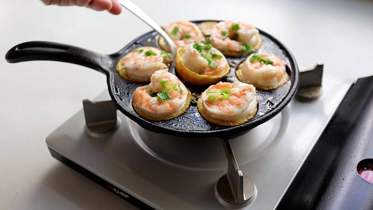 Ilustrasi banh khot, kue kecil khas Vietnam berbentuk bulat dengan topping udang dan daun bawang di atas wajan.
