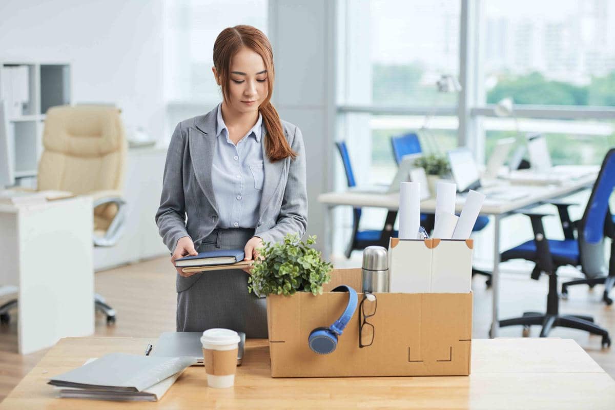 Seorang perempuan Asia berdiri di kantor dekat meja sambil membawa kotak kardus berisi barang pribadi saat resign dari pekerjaan.