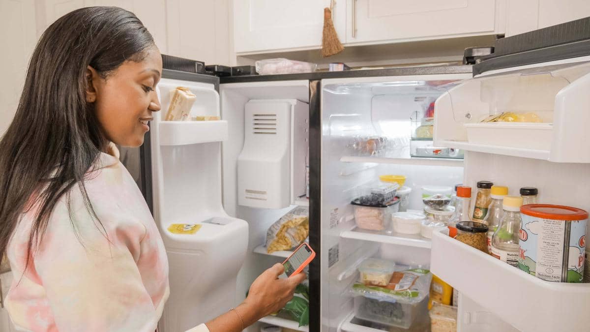 Seorang perempuan memeriksa isi kulkas terbuka sambil memegang wadah makanan di dapur rumah yang terang.